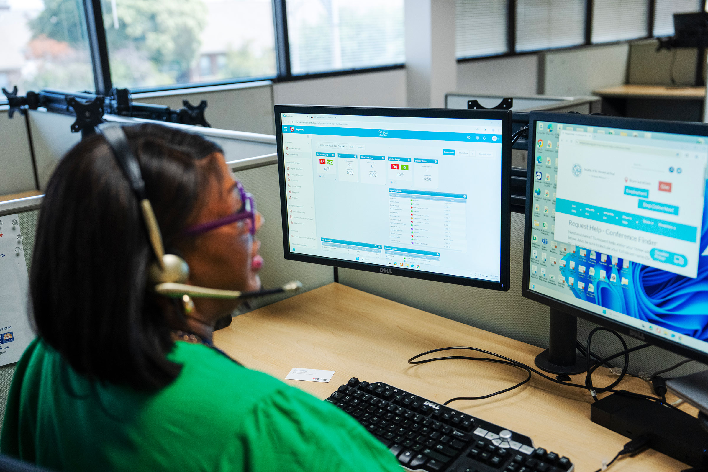 A woman working in a call center wears a headset and looks at two screens.