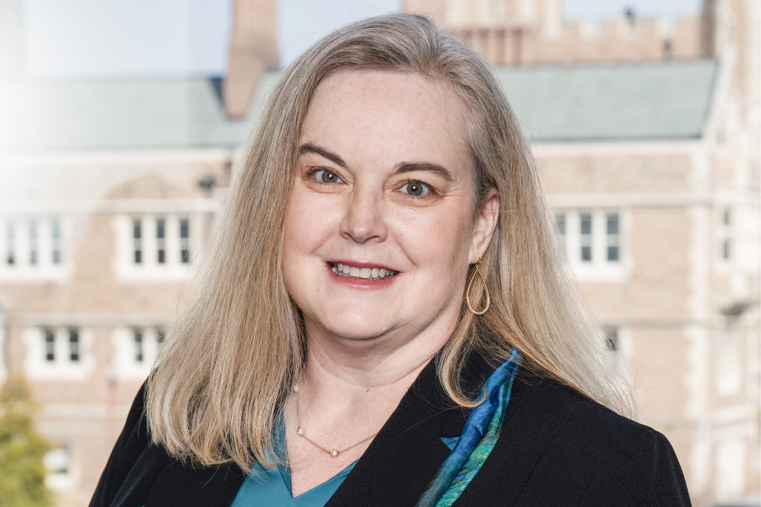 Tracy Bastain, a faculty member at the School of Public Health, is pictured wearing a black and blue top and smiling in front of Brookings Hall at WashU.