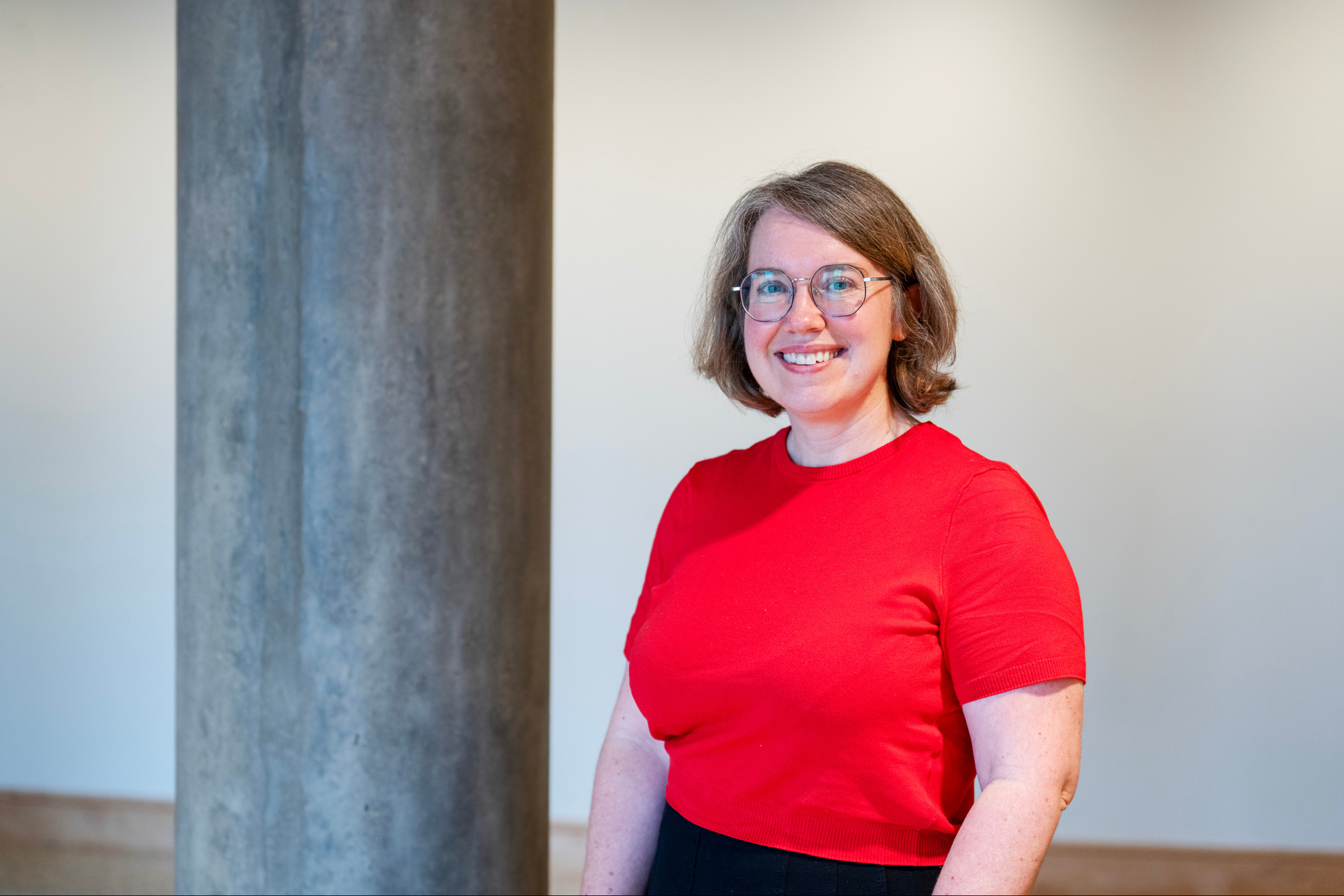 Susan Vorkoper, a staff member at the School of Pubic Health, poses with a smile wearing a red shirt against a plain white wall and a gray pillar.