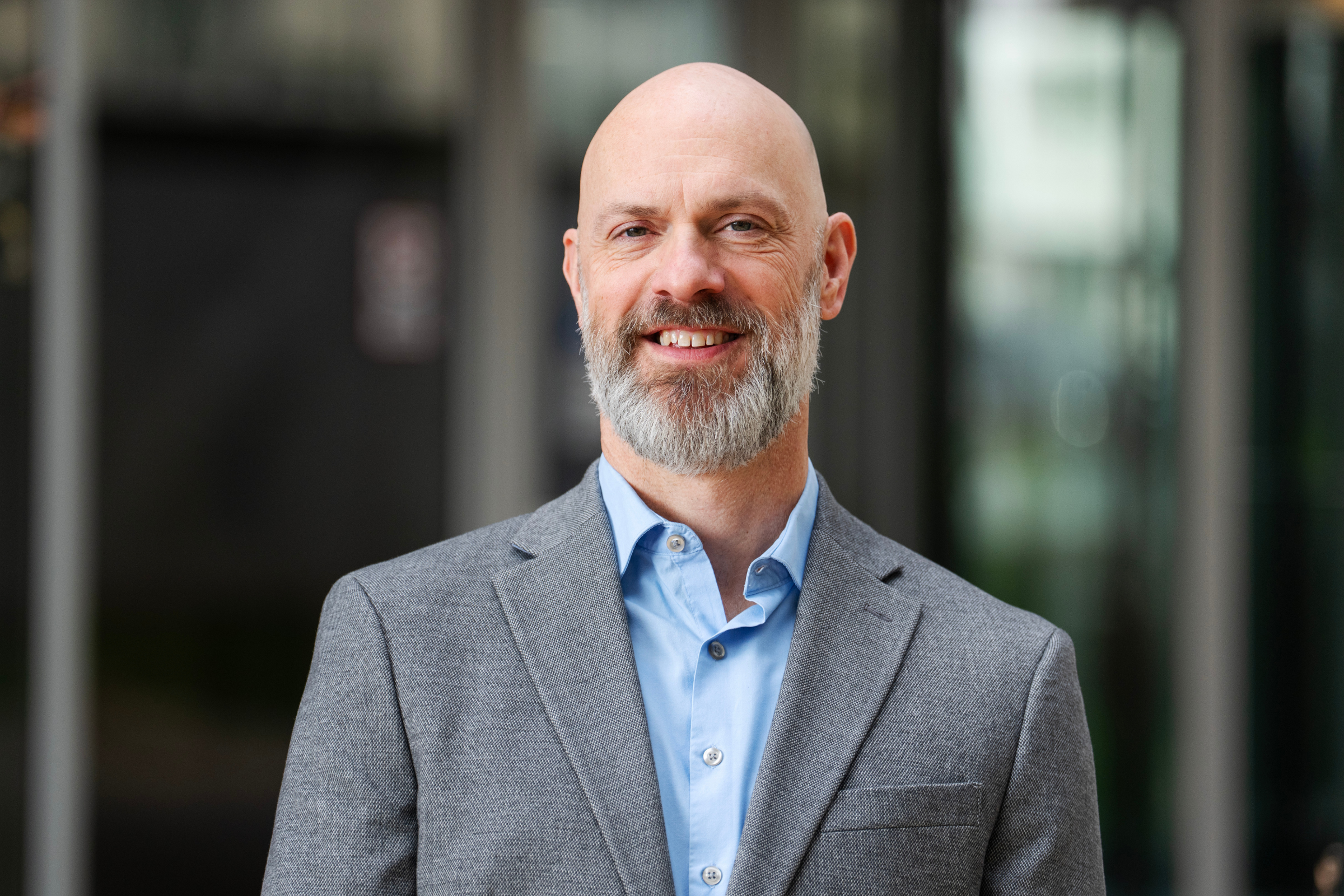 ben cooper, director of data management and analytics, stands in front of a blurred background wearing a gray blazer and blue collared button-up.