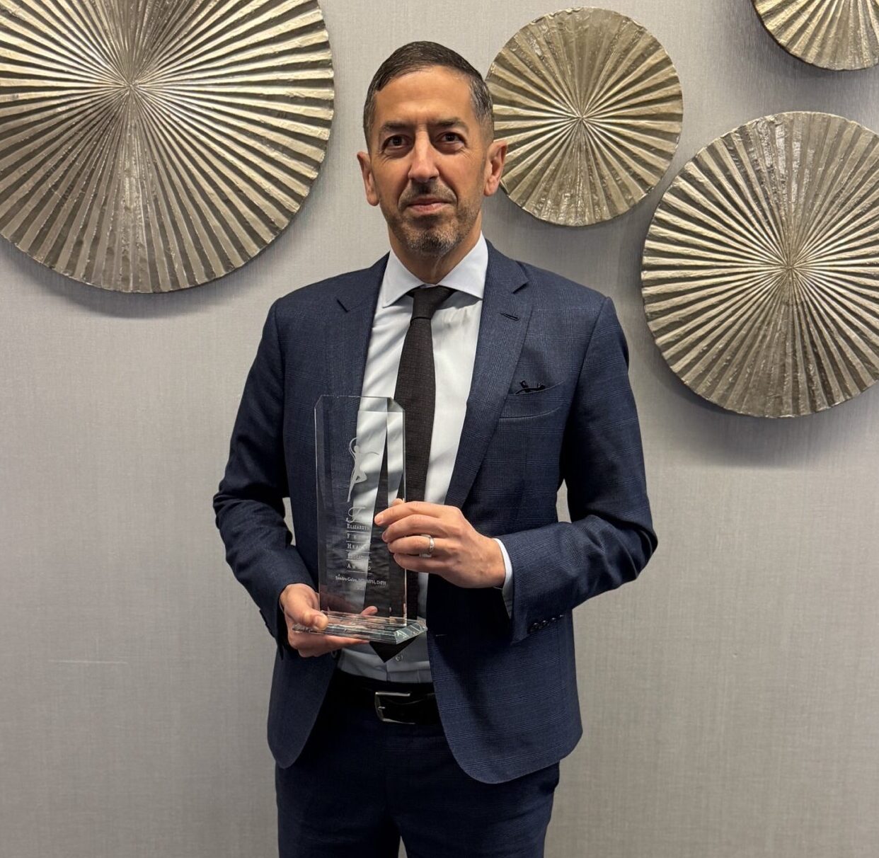 Dean Sandro Galea holding a crystal-like award he had just won; walls behind him are decorated with gold, textured circles