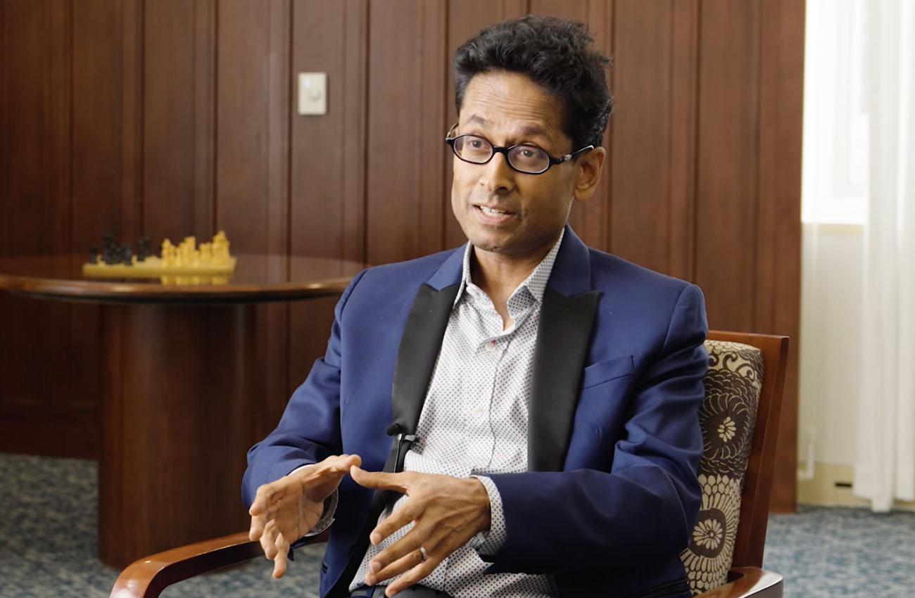 Man named Kumanan Rasanathan sitting in a chair in a nice room, answering questions while being interviewed by someone not in the image. He is wearing a nice jacket and using his hands to make expressions