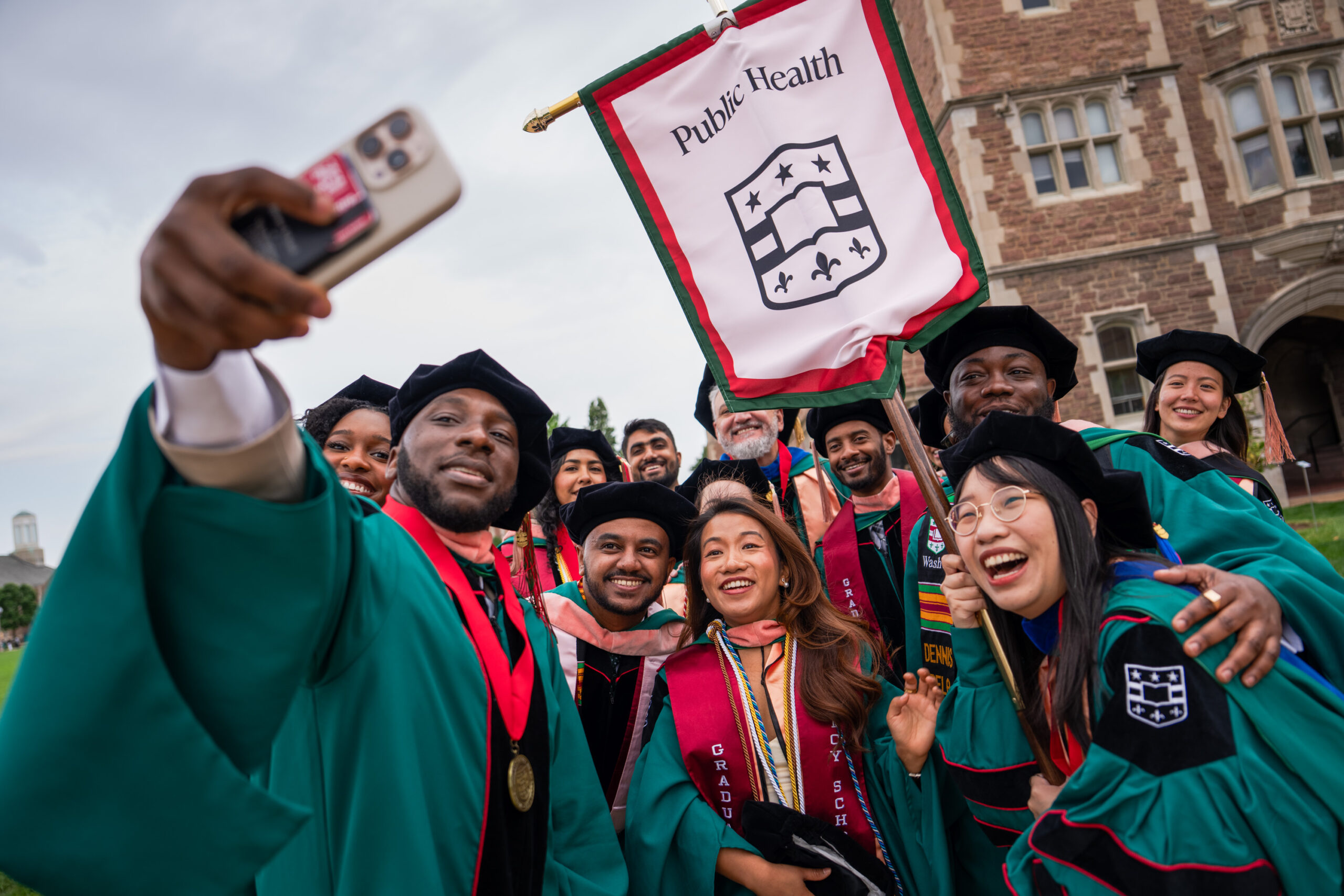 Graduation day photo showing happy students in graduation gowns, taking a photo, holding a "Public Health" flag